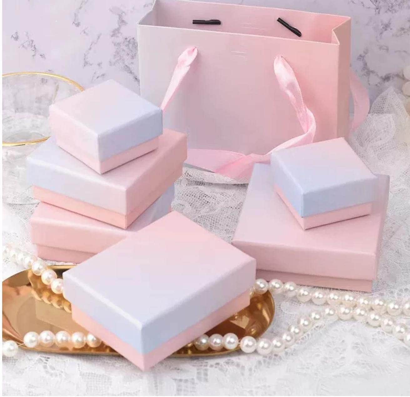 a pink gift bag, a set of pink and blue jewelry boxes, and a gold tray with pearls on a lace tablecloth.